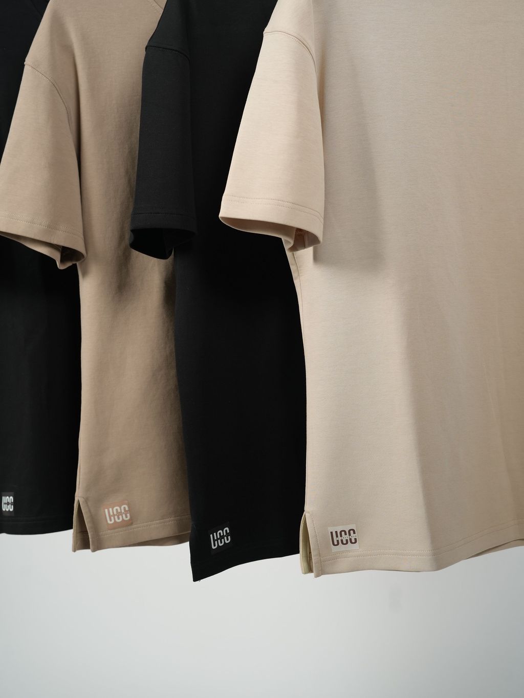 Two beige shirts and one black shirt hanging side by side on a white background